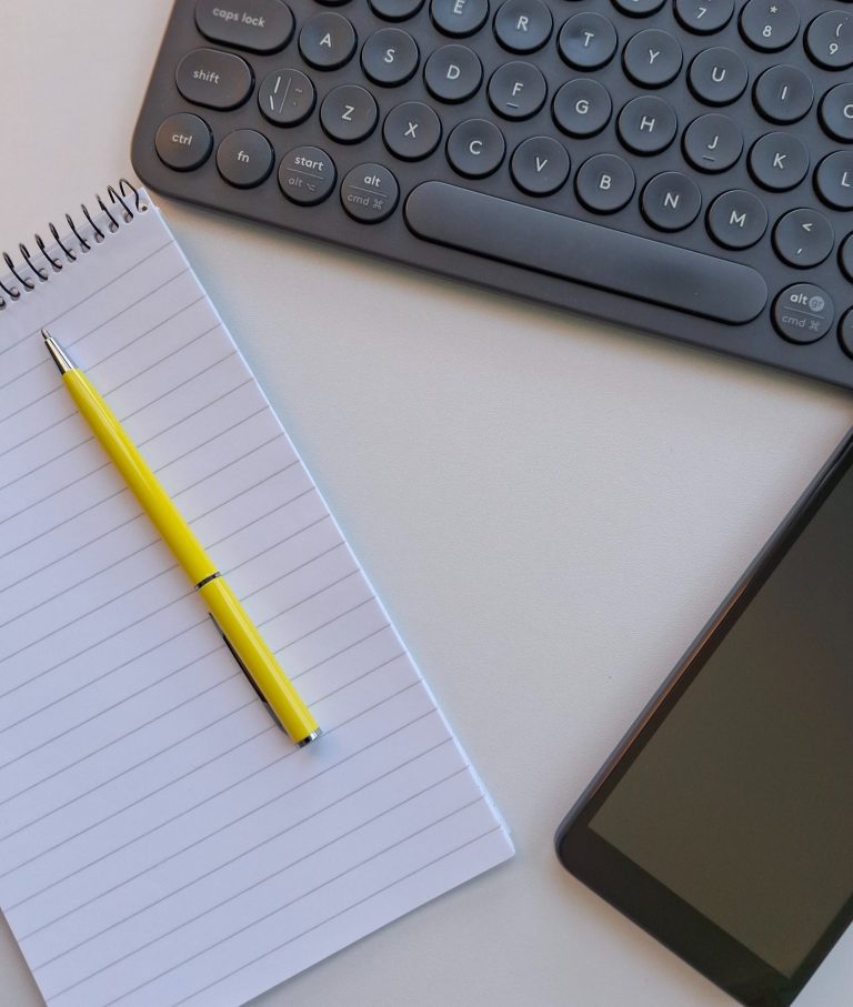 Pen, paper, mobile phone and keyboard A black keyboard with round keys, a spiral-bound notebook with a yellow pen resting on it, and a black smartphone on a white surface.