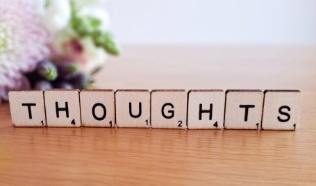 The word 'thoughts' spelled out in Scrabble tiles, placed on a surface with a softly blurred background of colourful flowers.