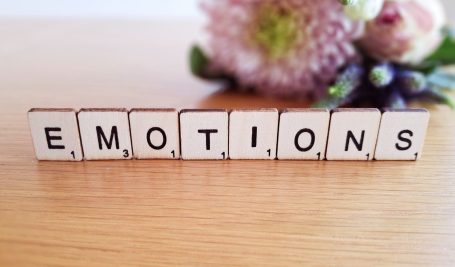 The word 'emotions' spelled out in Scrabble tiles, placed on a surface with a softly blurred background of colourful flowers.