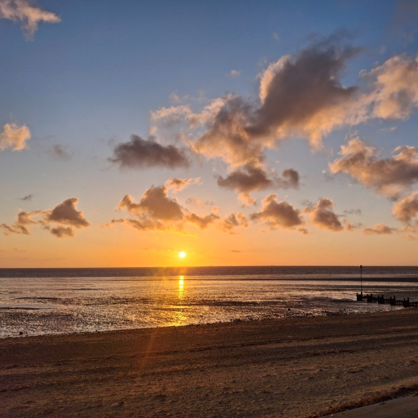 A serene beach at sunset with golden light reflecting on wet sand and water, scattered clouds, and an open horizon under a glowing sky.