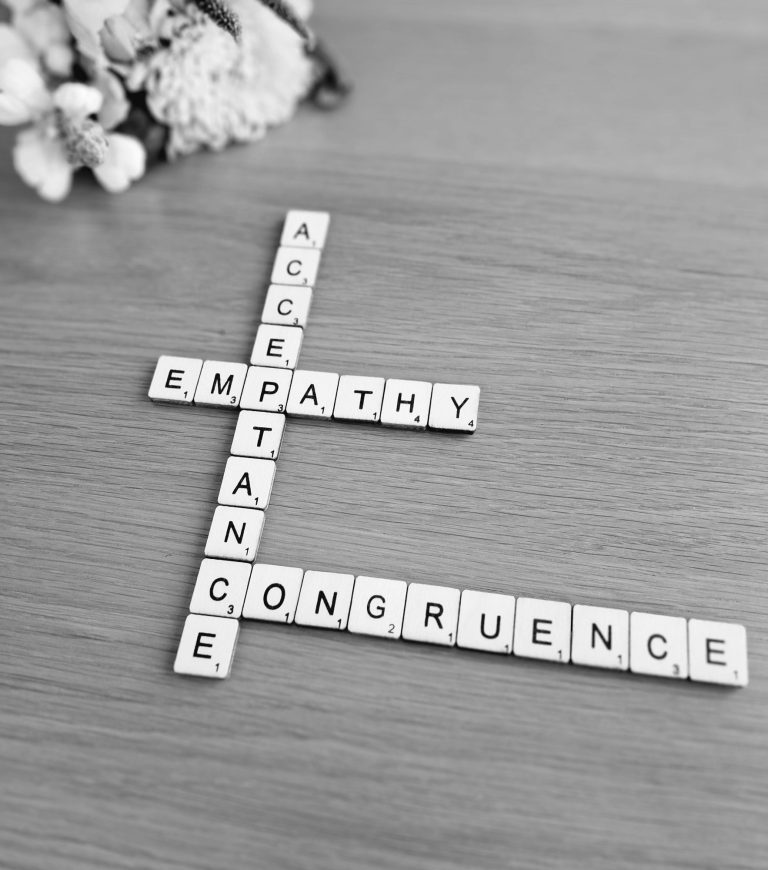Greyscale image Scrabble tiles spelling 'acceptance,' 'empathy' and 'congruence' on a surface, with blurred flowers in the top left corner.