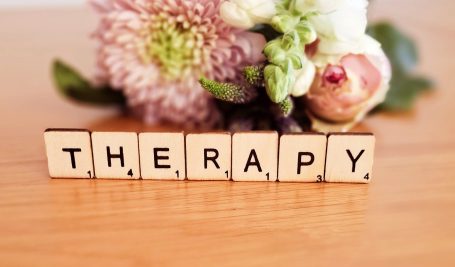 The word 'therapy' spelled out in Scrabble tiles, placed on a surface with a softly blurred background of colourful flowers.