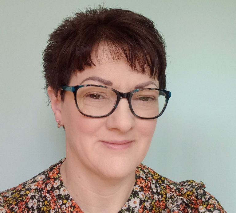 Natalie Wenham A selfie of Natalie with short, spiky brown hair, brown eyes, teal-coloured glasses, flowery top and pale blue background.