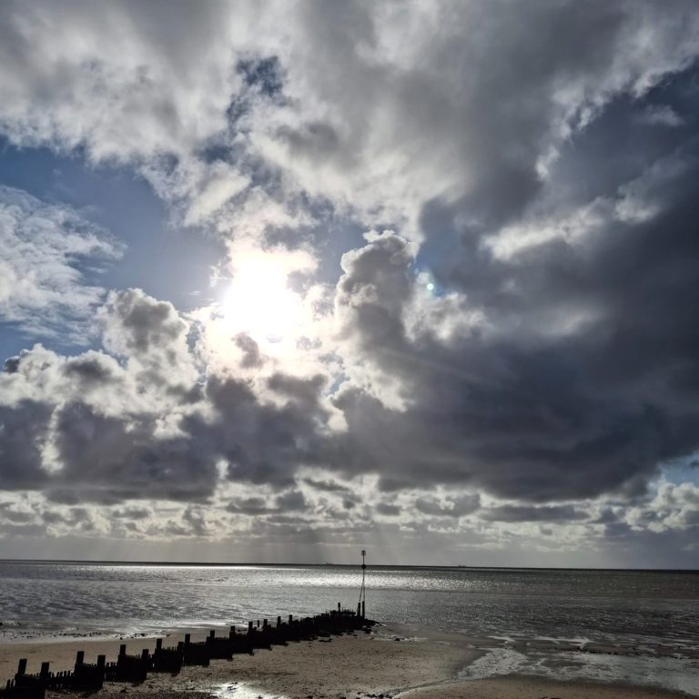 A dramatic beach scene with dark clouds, sunlight breaking through, wet sand, scattered rocks, and a wooden groyne stretching into the water